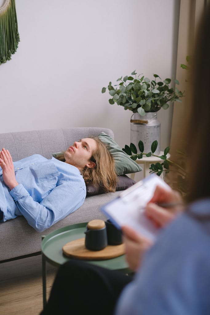 A person in therapy lying on a sofa with a therapist taking notes nearby.