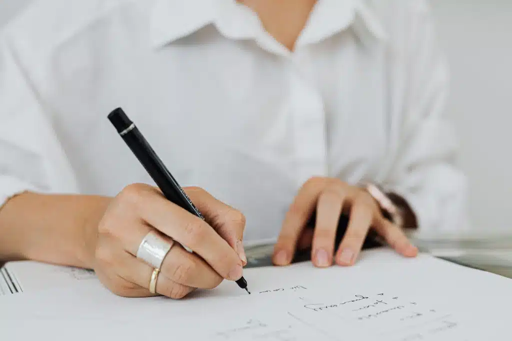 A person in a white shirt is writing on a paper with a black pen. The focus is on the hand adorned with a large silver ring, capturing a sense of concentration.
