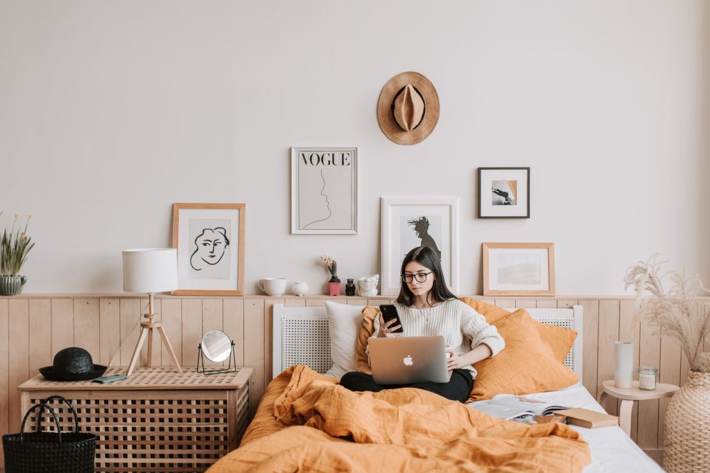 A young woman wearing glasses and a sweater sits on a bed with orange bedding, using a laptop on her lap while looking at her smartphone.