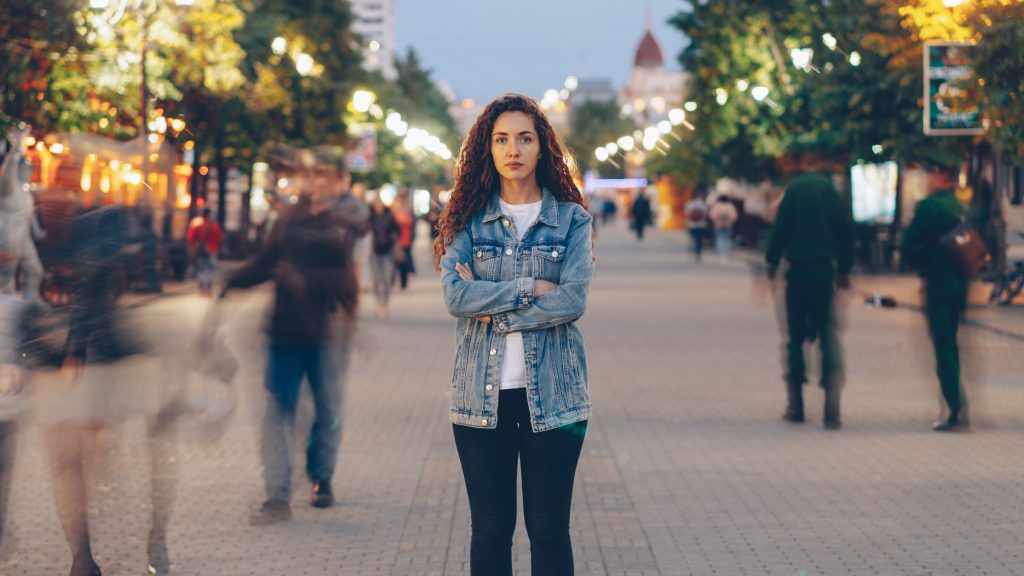 A young woman with curly hair wearing a denim jacket standing still on a busy pedestrian street at dusk with arms crossed, as crowds of people blur by in the background.