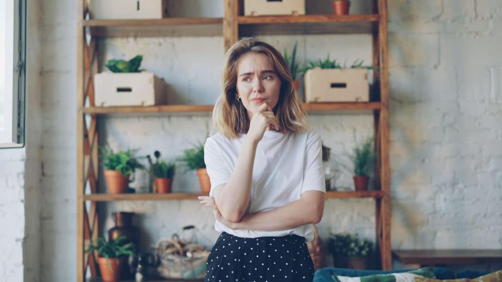 A thoughtful young woman standing indoors with her hand on her chin, with a rustic wooden shelving unit containing potted plants and boxes in the background.